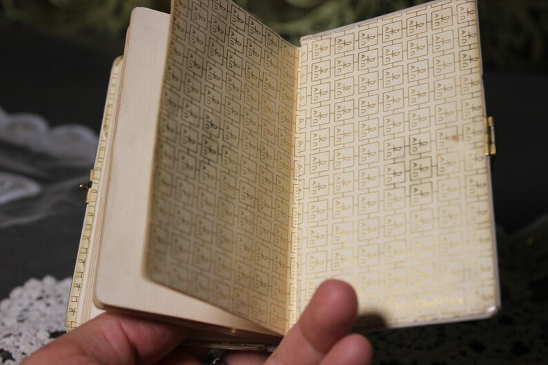 May include: A small, vintage notebook with a cream-colored cover and pages. The interior pages feature a repeating gold pattern. The notebook is open, revealing the patterned interior. A gold clasp secures the notebook.