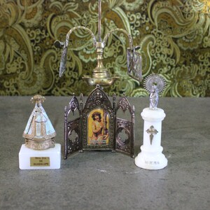 May include: A set of three small religious figurines on a marble surface. The figurines are made of metal and feature a silver finish. The figurine on the left is of the Virgin of Guadalupe, the middle figurine is of Jesus Christ, and the figurine on the right is of a saint with a halo. The figurines are all on small marble bases.