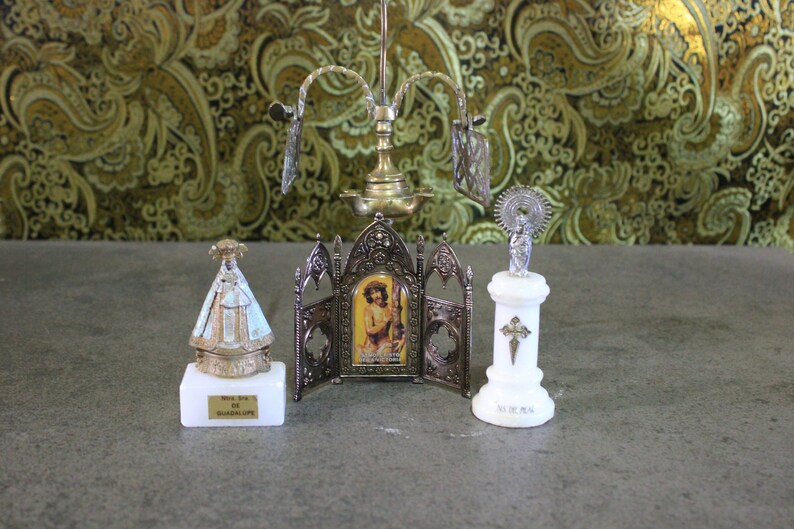 May include: A set of three small religious figurines on a marble surface. The figurines are made of metal and feature a silver finish. The figurine on the left is of the Virgin of Guadalupe, the middle figurine is of Jesus Christ, and the figurine on the right is of a saint with a halo. The figurines are all on small marble bases.