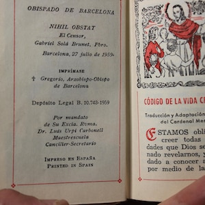 May include: Open book with text in Spanish, including "OBISPADO DE BARCELONA" and "C&Oacute;DIGO DE LA VIDA CRISTIANA." The pages have red borders and a small illustration. The book is held open by a hand.