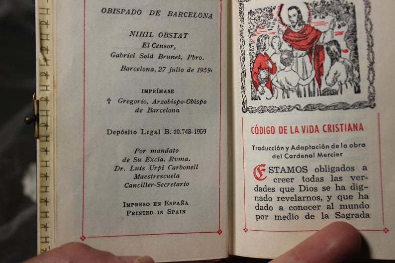May include: Open book with text in Spanish, including "OBISPADO DE BARCELONA" and "C&Oacute;DIGO DE LA VIDA CRISTIANA." The pages have red borders and a small illustration. The book is held open by a hand.