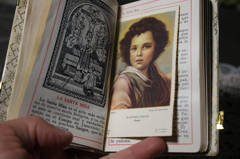 May include: An open, antique book with gold-colored edges and a decorative cover. The left page features a black and white illustration and text in Spanish. The right page displays a color image of a young person. The text "EL DIVINO PASTOR" is visible.