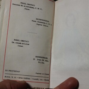 May include: Open book with text in Spanish. The left page has text in red and black ink, including "NIHIL OBSTAT" and names. The right page is blank with a faint image. The book's edge has a decorative gold pattern.