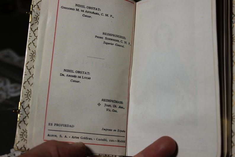 May include: Open book with text in Spanish. The left page has text in red and black ink, including "NIHIL OBSTAT" and names. The right page is blank with a faint image. The book's edge has a decorative gold pattern.