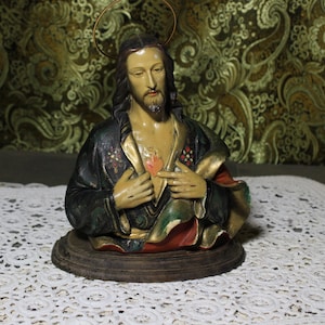 Puede incluir: Busto de Jesucristo con aureola, vestido con una túnica oscura con detalles dorados y un corazón rojo en el pecho. La estatua está montada sobre una base de madera y colocada sobre un fondo estampado.