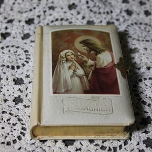 May include: A small, vintage book with a religious illustration on the cover. The image depicts a young girl in a white veil receiving communion from Jesus. The book has a gold clasp and the word "COMUNION" is visible.