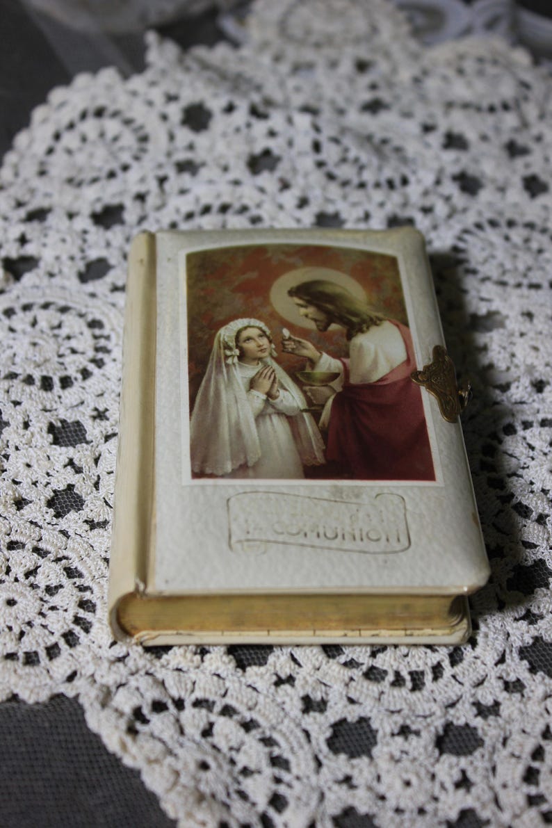 May include: A small, vintage book with a religious illustration on the cover. The image depicts a young girl in a white veil receiving communion from Jesus. The book has a gold clasp and the word "COMUNION" is visible.