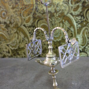 May include: A brass oil lamp with a decorative design. The lamp has two side arms with ornate metal panels and a central spout. The lamp sits on a round base.