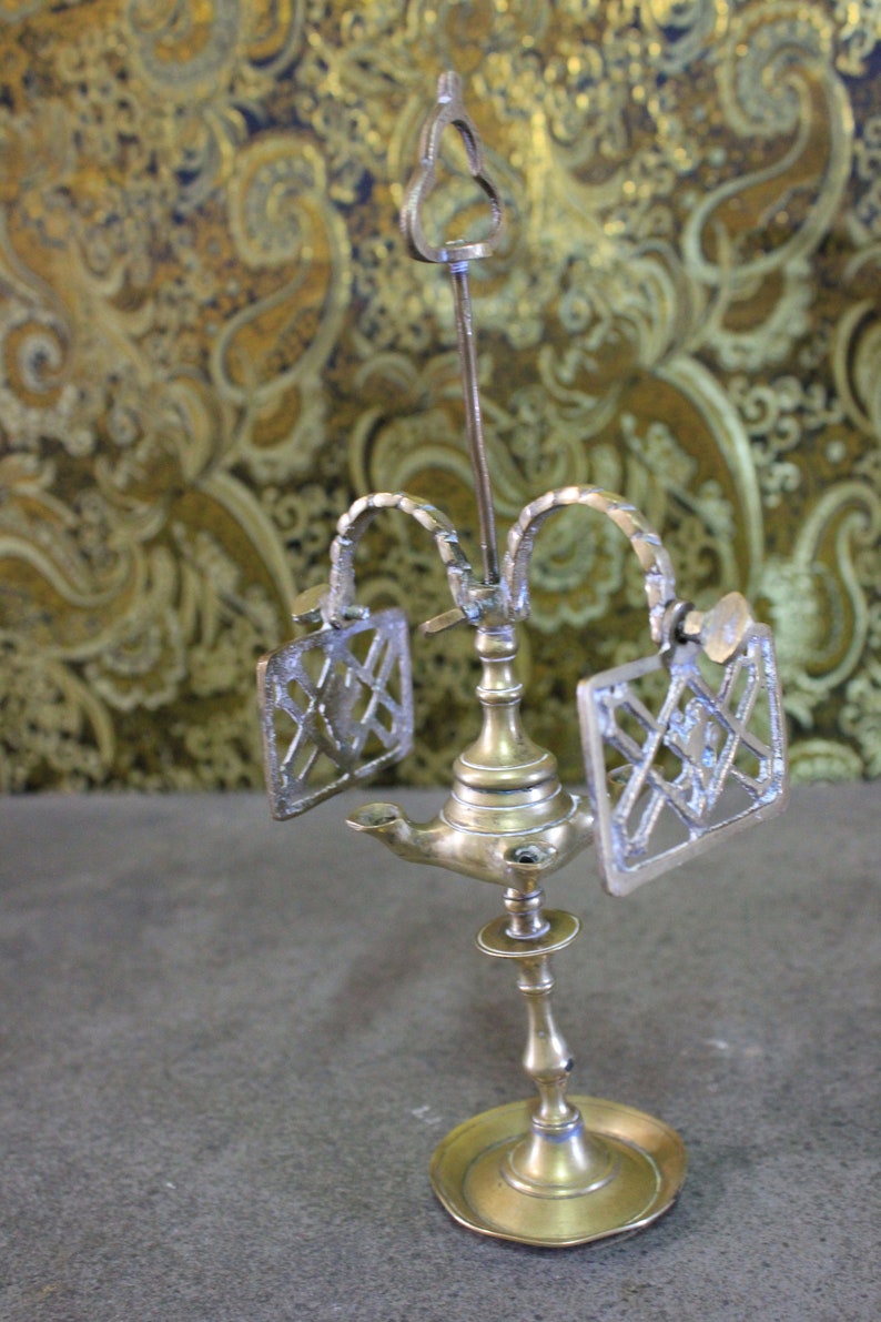 May include: A brass oil lamp with a decorative design. The lamp has two side arms with ornate metal panels and a central spout. The lamp sits on a round base.