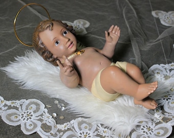 May include: A ceramic figurine of a baby Jesus wearing a gold halo and a white and yellow robe. The figurine is lying on a white fluffy surface with a white lace trim.