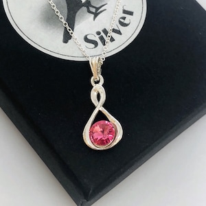 May include: A silver infinity necklace with a pink crystal pendant. The necklace is on a black background.