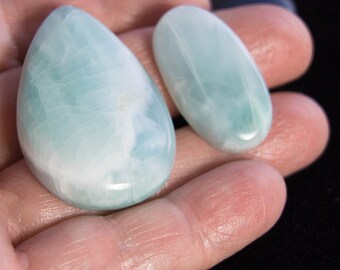 May include: Two polished, smooth, light blue and white stones. One stone is shaped like a teardrop, while the other is an elongated oval. Both stones feature a marbled, cloudy texture with varying shades of pale blue and white throughout the surface.