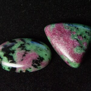 Peut inclure: Deux pierres précieuses polies présentant un motif tacheté de magenta vibrant, de vert forêt profond et d'inclusions noires. Une pierre est en forme d'ovale lisse, tandis que l'autre est un triangle arrondi. Les deux ont une surface brillante.