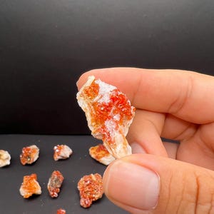 Può includere: Una collezione di piccoli gruppi di cristalli di colore arancione e bianco. I cristalli sono disposti su una superficie nera.