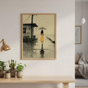 May include: Framed art print depicting a person in a yellow dress walking with an umbrella on a rainy day. The artwork features a grayscale building and a reflective surface, with a light wood frame.