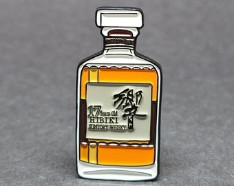 Könnte beinhalten: Eine goldene und weiße Emaille-Anstecknadel in Form einer Flasche Hibiki Suntory Whisky. Die Flasche trägt ein Etikett mit der Aufschrift "17 Years Old Hibiki Suntory Whisky" und zeigt japanische Schriftzeichen.