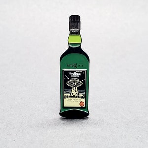 Puede incluir: Un pin de esmalte verde con forma de botella de whisky con una etiqueta negra. La etiqueta dice "ESTD 1815" y "The Ultimate" con un gráfico de un OVNI y el texto "Supernova" encima.