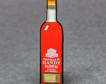 May include: A pin shaped like a bottle of Thomas H. Handy Sazerac Straight Rye Whiskey. The bottle is red with a white label featuring the brand name and a building illustration. The top of the bottle is gold.