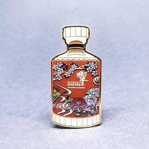 May include: Gold-colored enamel pin shaped like a bottle of Hibiki Suntory Whisky. The bottle is decorated with a red and white floral design and the text "Hibiki Suntory Whisky Japanese Harmony".