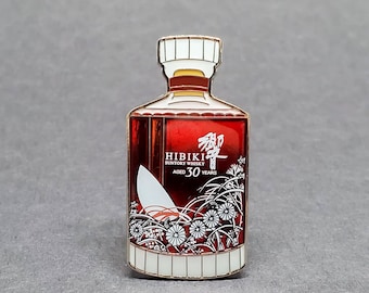 May include: A gold-colored enamel pin shaped like a bottle of Hibiki Suntory Whisky, aged 30 years. The bottle is red with white lettering and a white floral design.