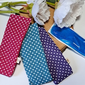 Könnte beinhalten: Drei Augenmasken-Beutel mit weißen Punkten auf rotem, türkisfarbenem und violettem Hintergrund. Die Beutel liegen auf einer Holzoberfläche mit einem blauen Kühlkissen.