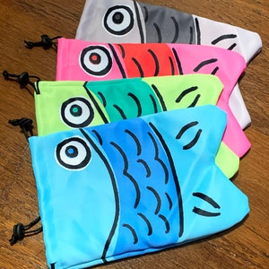 May include: Five colorful drawstring bags with fish designs. The bags are made of fabric and have a drawstring closure. The fish designs are different colors and have black outlines. The bags are stacked on top of each other.