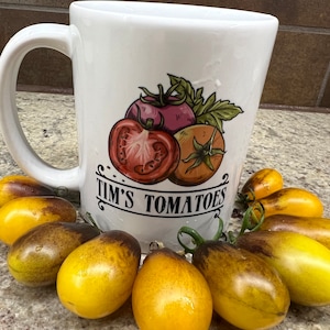 May include: White ceramic mug with a drawing of three tomatoes and the text "Tim's Tomatoes".  The mug is surrounded by a ring of small yellow tomatoes with brown stripes.