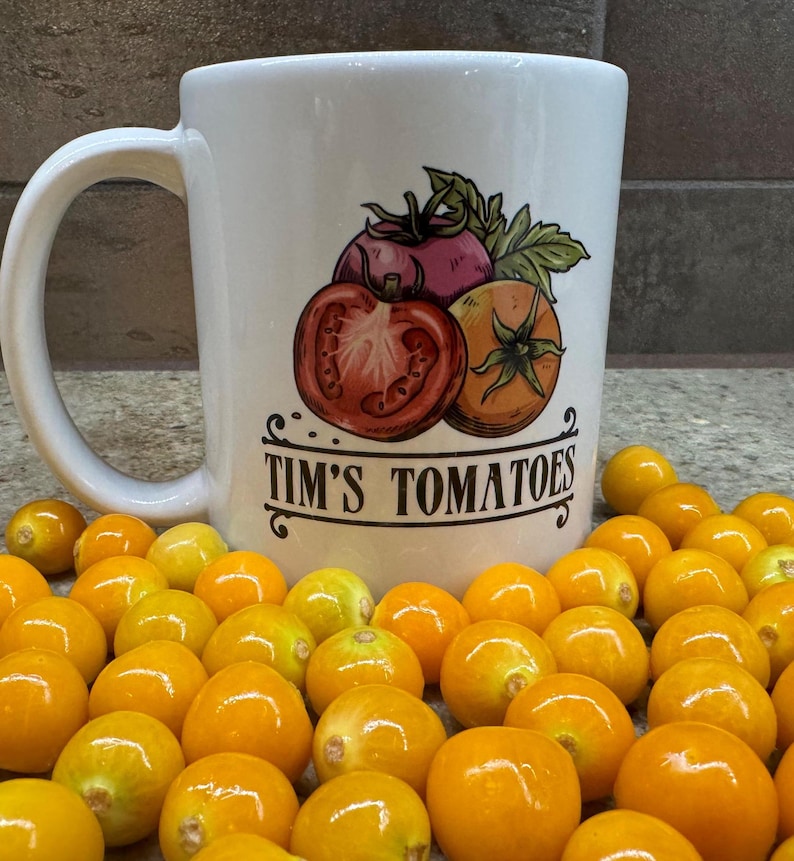 May include: White ceramic mug featuring a red, purple, and orange tomato illustration with the text "Tim's Tomatoes". The mug is surrounded by small, bright yellow, round fruits.