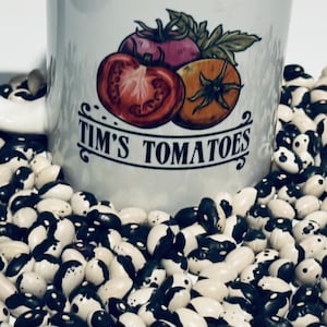 May include: A white ceramic mug with the text "TIM'S TOMATOES" and a colorful illustration of tomatoes. The mug is surrounded by a pile of black and white beans.