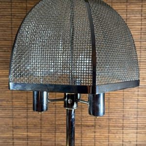 May include: A vintage chrome table lamp with a mesh dome shade. The lamp has a polished chrome base and three cylindrical light fixtures. The shade is a fine metal mesh, giving the lamp a retro aesthetic.