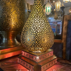 May include: A gold Moroccan table lamp with a teardrop-shaped shade and intricate cut-out designs. The lamp has a three-tiered base with a red light shining through the cut-outs.