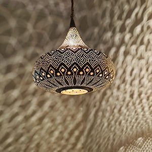May include: A decorative black metal pendant light with a patterned design of small holes. The light is suspended from a chain and casts intricate shadows on the wall. The light has a teardrop shape.
