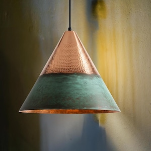 Puede incluir: Una lámpara colgante de cobre con forma cónica. La parte superior tiene una textura martillada y un color cobre cálido, mientras que la sección inferior es de color verde desgastado. Cuelga de un cable negro.