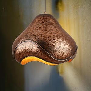 Pode incluir: Uma luminária pendente de cor cobre com uma textura martelada. A luz tem um formato curvo único com um brilho dourado e quente que emana da parte inferior. A luminária pende de um cabo preto, mostrando um design moderno.