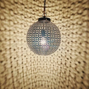 以下が含まれることがあります： チェーンから吊り下げられた、模様入りの金属シェードが付いた球形のペンダントライト。シェードのデザインは、周囲の表面に複雑な影を落とします。照明器具が点灯し、温かい雰囲気を作り出しています。