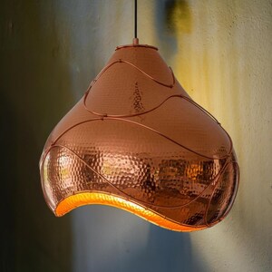 May include: Copper pendant light with a hammered texture and a unique, curved shape. The warm, metallic finish and decorative wire accents create a stylish, modern look. The light fixture hangs from a black cord.