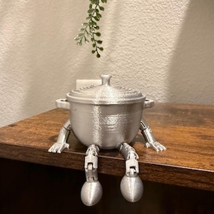 May include: A silver-colored, 3D-printed pot with a lid, handles, and articulated legs and feet. The pot is sitting on a wooden surface. A sprig of green foliage hangs in the background.