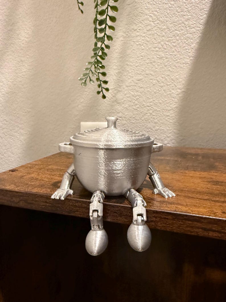 May include: A silver-colored, 3D-printed pot with a lid, handles, and articulated legs and feet. The pot is sitting on a wooden surface. A sprig of green foliage hangs in the background.