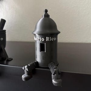 May include: A black decorative figurine of a fort with the text "Puerto Rico" on the front. The figurine has legs and is sitting on a black surface. The fort has a small window and a rounded top.