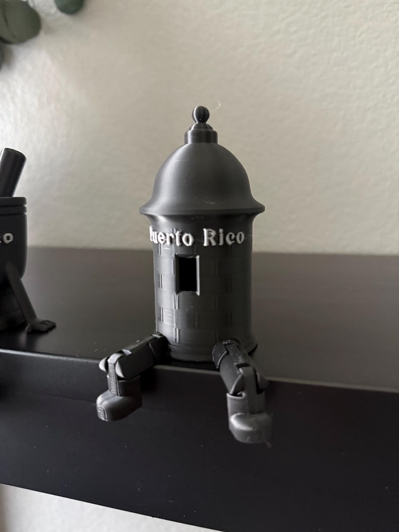 May include: A black decorative figurine of a fort with the text "Puerto Rico" on the front. The figurine has legs and is sitting on a black surface. The fort has a small window and a rounded top.