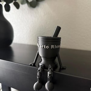 May include: Black mortar and pestle with the words "Puerto Rico" in silver lettering. The mortar has arms and legs, appearing to sit on a black surface. A black vase and greenery are in the background.