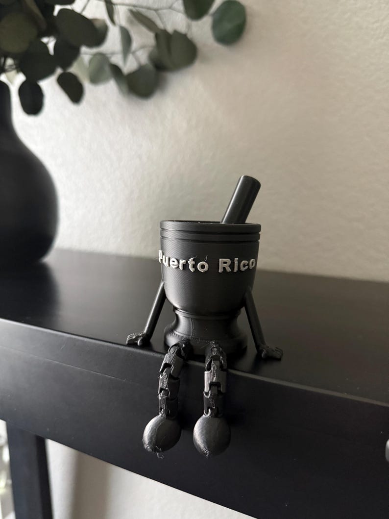 May include: Black mortar and pestle with the words "Puerto Rico" in silver lettering. The mortar has arms and legs, appearing to sit on a black surface. A black vase and greenery are in the background.