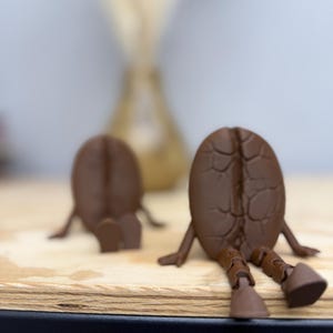May include: Two brown coffee bean figurines with legs and arms, sitting on a wooden surface. The figurines have a textured surface, and are in a relaxed pose. The background is blurred, with a light-colored vase.
