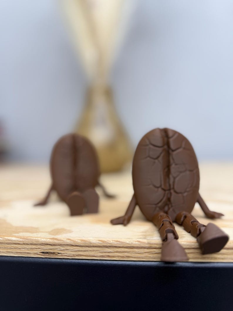 May include: Two brown coffee bean figurines with legs and arms, sitting on a wooden surface. The figurines have a textured surface, and are in a relaxed pose. The background is blurred, with a light-colored vase.