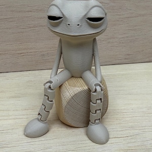 May include: A gray, articulated frog figurine with closed eyes, seated on a wooden, rounded base. The frog has jointed legs and arms, and a relaxed expression. The background is a light brown wooden surface.