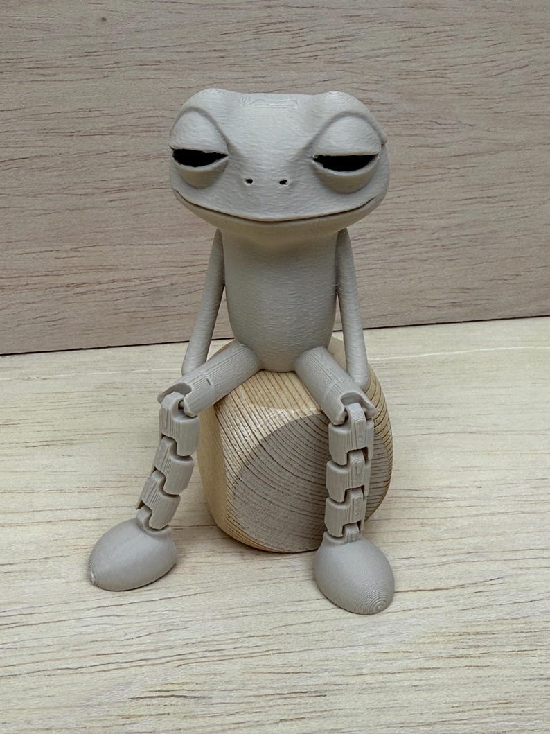 May include: A gray, articulated frog figurine with closed eyes, seated on a wooden, rounded base. The frog has jointed legs and arms, and a relaxed expression. The background is a light brown wooden surface.