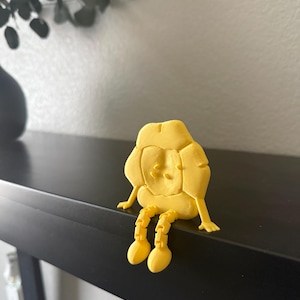 May include: A yellow, 3D-printed figurine of a seated character with a rounded head and segmented legs. The figure has a simple face and is sitting on a black surface. The background includes a wall and a dark vase with greenery.