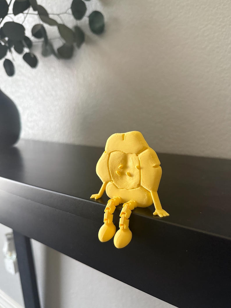 May include: A yellow, 3D-printed figurine of a seated character with a rounded head and segmented legs. The figure has a simple face and is sitting on a black surface. The background includes a wall and a dark vase with greenery.