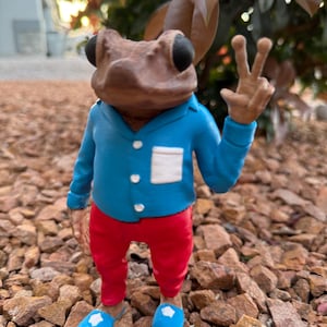 May include: A whimsical figurine of a frog wearing a blue jacket, red pants, and blue slippers. The frog is giving a peace sign with its right hand. The figurine is standing on a bed of small rocks, with a blurred background of greenery.
