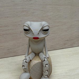 May include: A whimsical, gray frog figurine with articulated limbs and a bored expression. The frog is seated on a wooden, rounded base and has red lipstick. The frog's design is reminiscent of a puppet or toy.